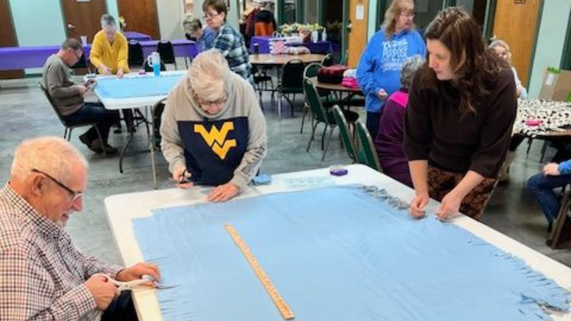 People gathered around a large blue quilt, measuring and arranging fabric pieces in a community room.