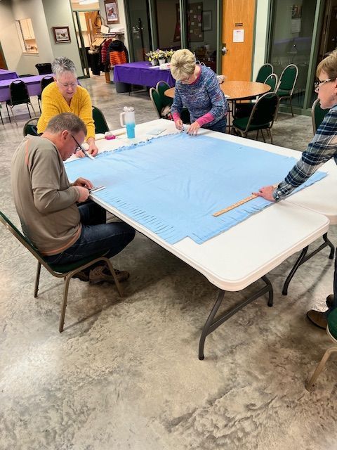 People gathered around a table folding a large blue cloth in a community room