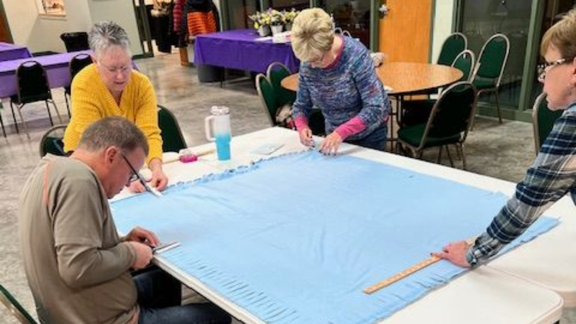 Four people gather around a large blue board, working together with markers in a community room.