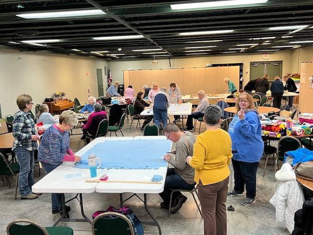 People gathered at tables in a busy community hall, chatting and browsing crafts and supplies.