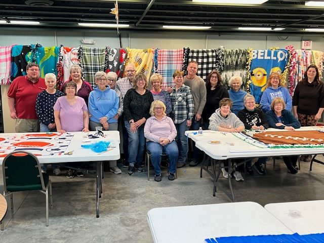 Group of people posing behind decorated tables at an indoor event with colorful quilts hanging behind them
