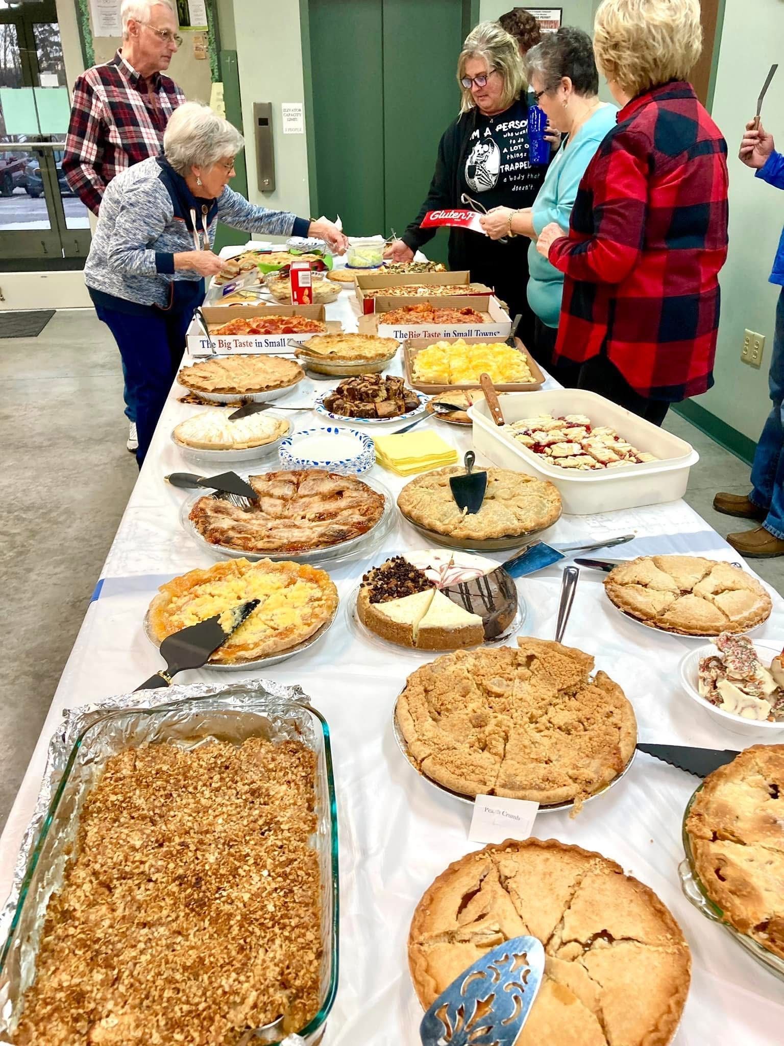 People serving and selecting homemade pies at a buffet table in a community hall