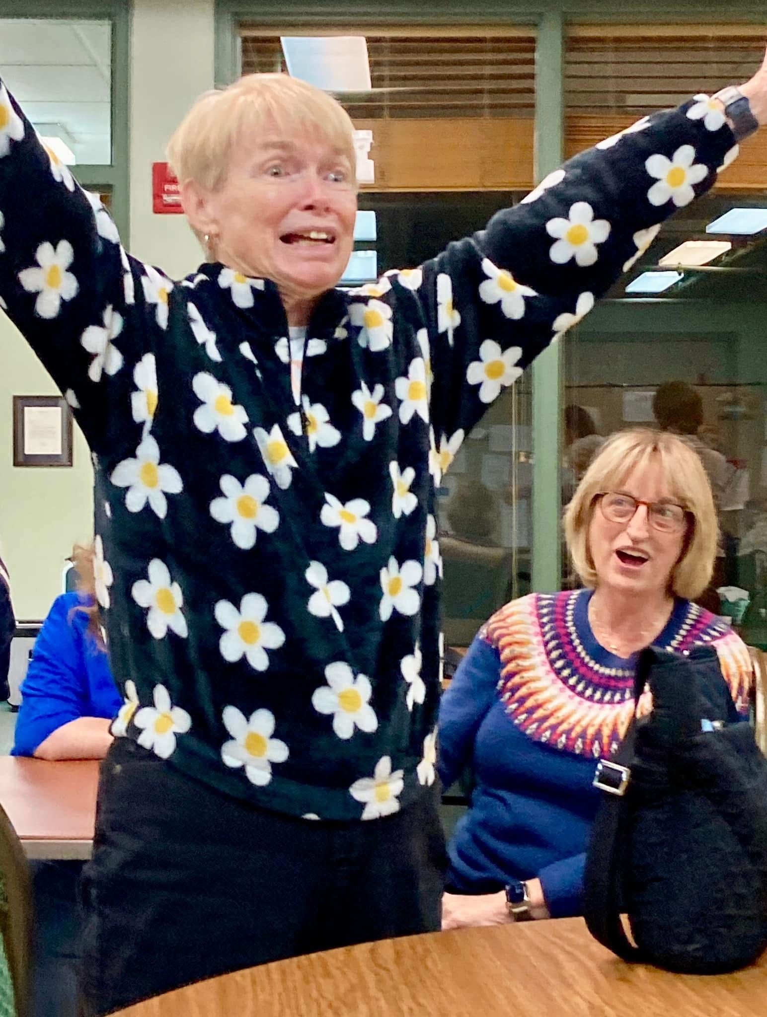 Person in a daisy sweater raises arms while others sit nearby in a room.
