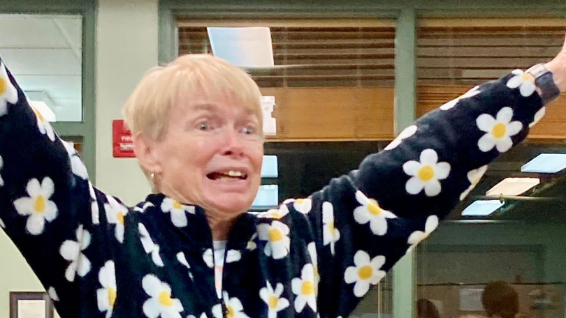 Person in a daisy-patterned sweater with arms raised, smiling indoors in front of windows.