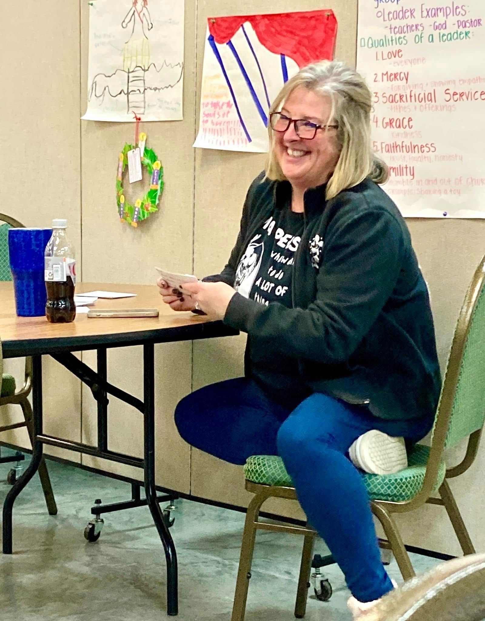 Woman smiling at a table in a classroom, holding a cup with posters on the wall behind her