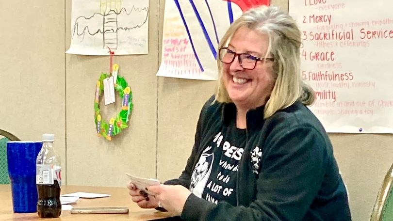 Woman smiling at a desk in a classroom, holding a phone with posters and a wreath behind her.