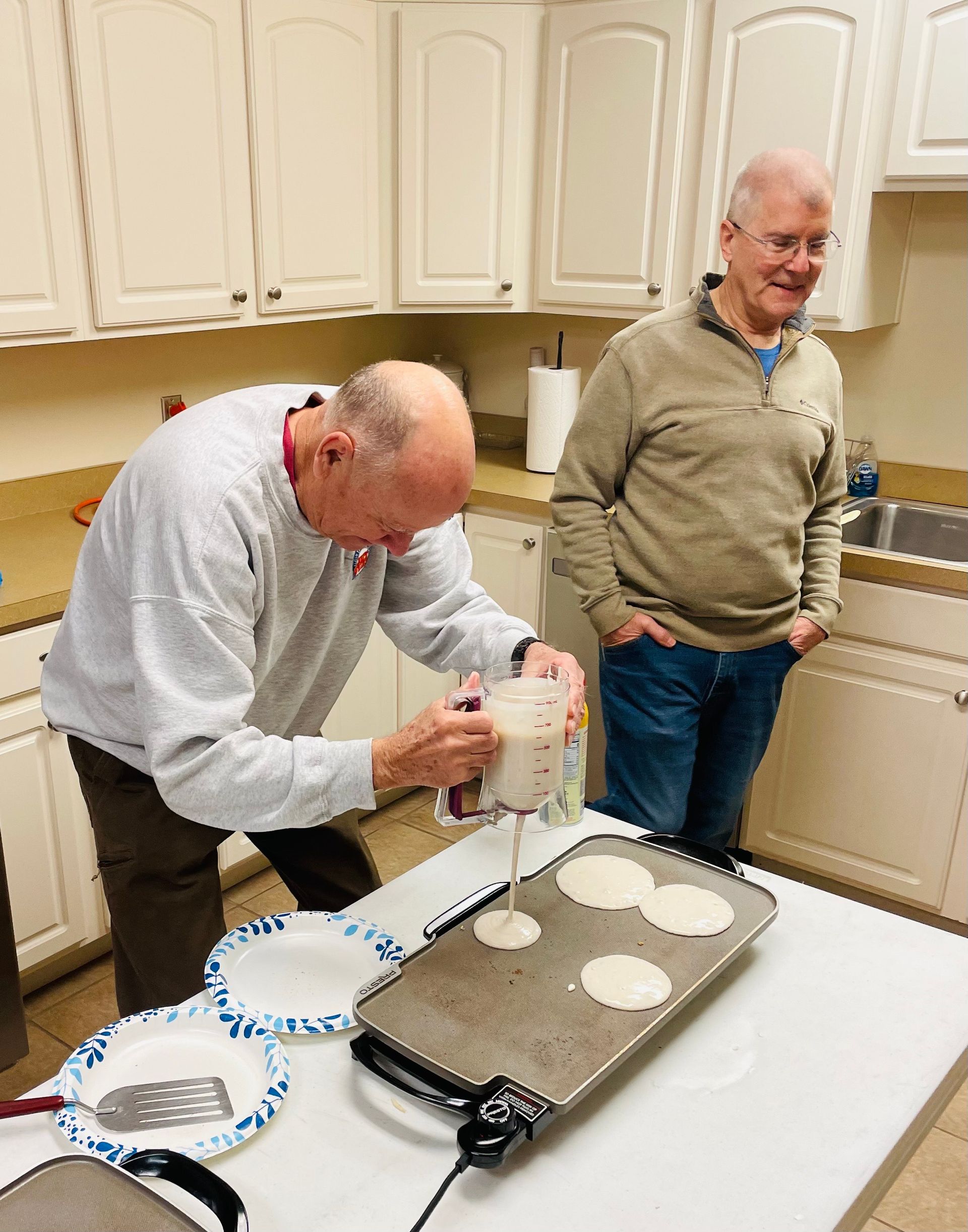 Two men in a kitchen prepare dough beside a griddle with pancakes and stacked plates.