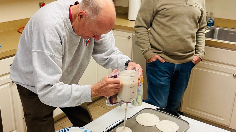 Man stirring batter in a mixing bowl at a kitchen counter while another person stands nearby