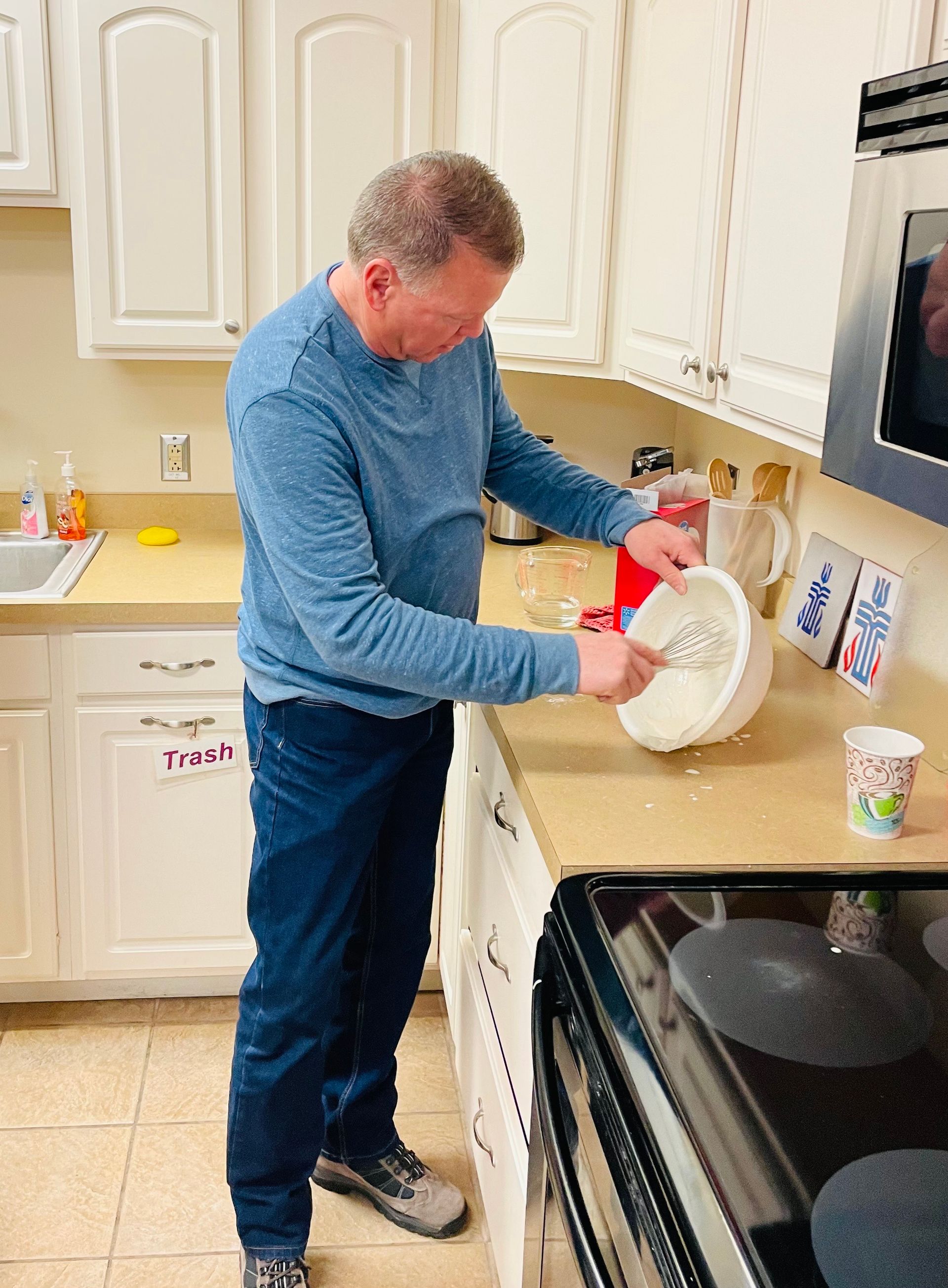 Man washing dishes at a kitchen sink beside a stove, holding a white plate and red dish soap.
