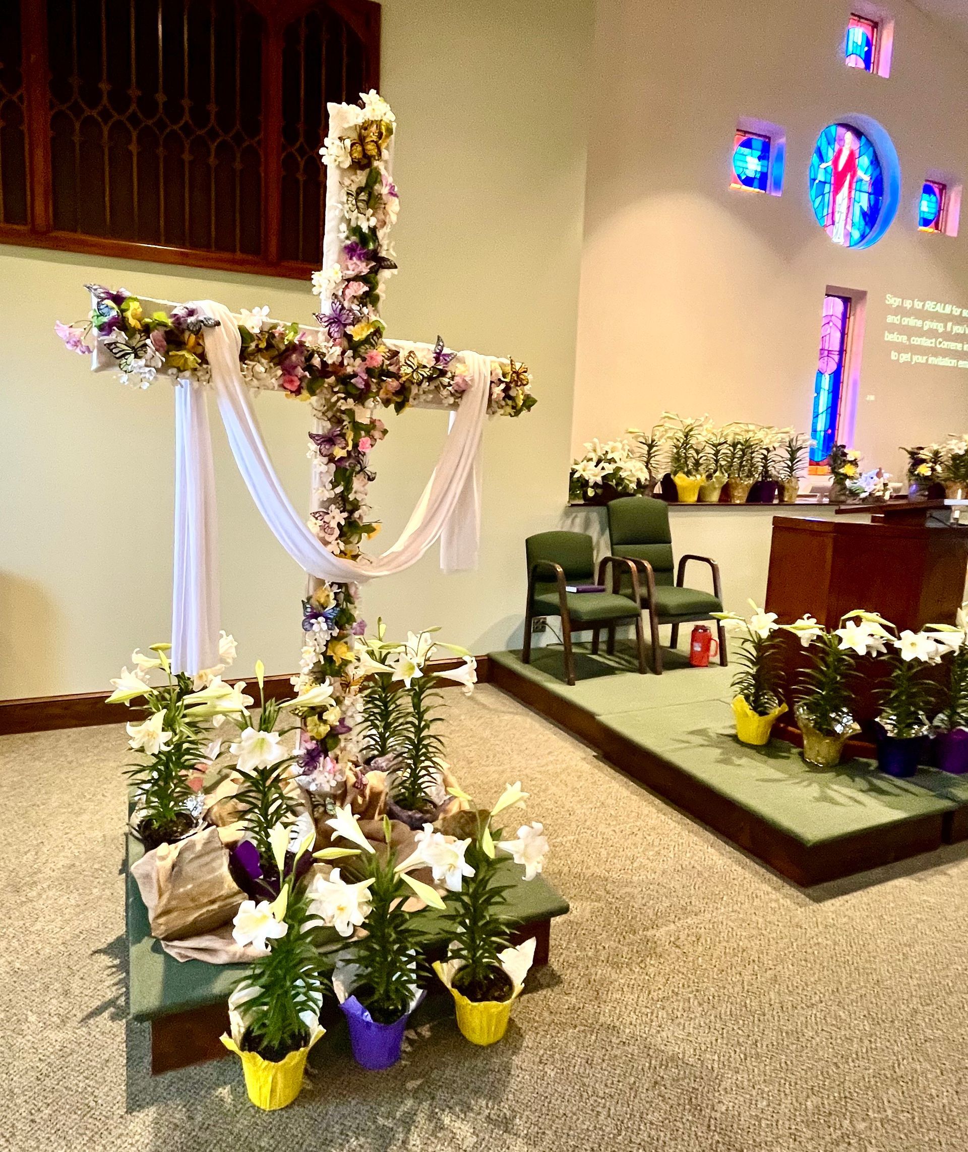 Flower-covered cross on a church stage with white drapery and potted plants around the altar