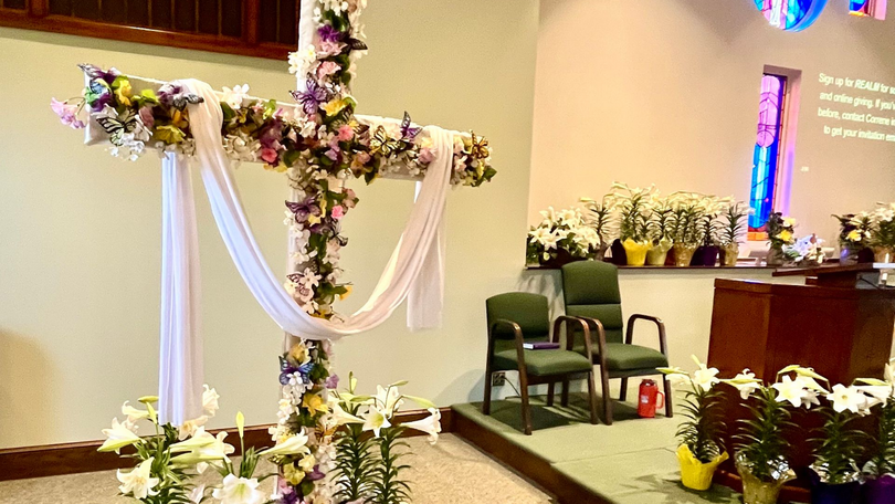 Flower-covered cross on a church altar with white drapery, chairs, and floral arrangements