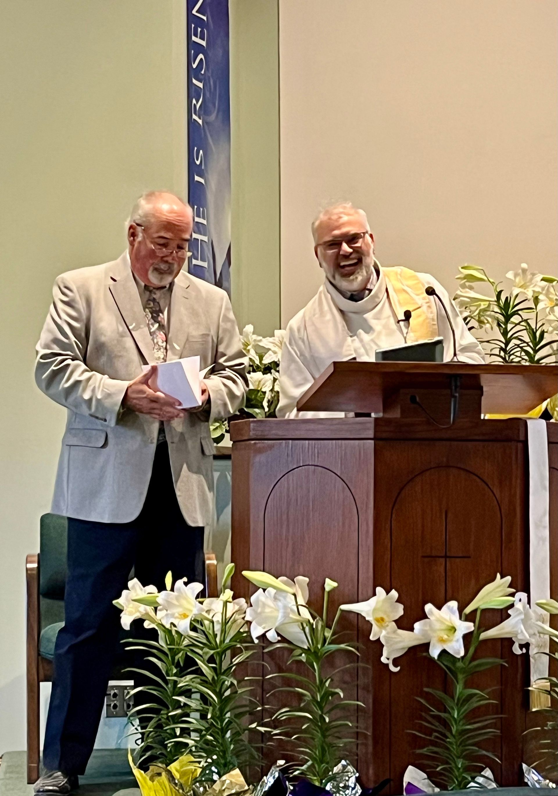 Two men at a church lectern with white flowers in front and a blue banner behind them