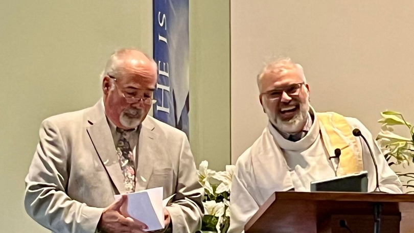 Two men speaking at a podium, one holding notes, in a conference setting with a banner behind them.