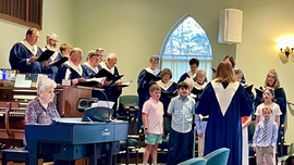 Choir and children sing in a church sanctuary during a performance, with a pianist at left and conductor at center.