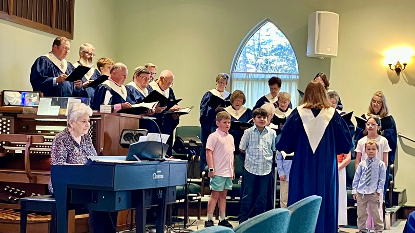 Choir singing in a church sanctuary, led by a robed conductor beside a piano and microphone stand.