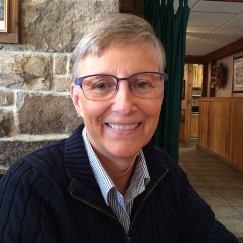 Smiling person wearing glasses and a dark sweater, seated indoors beside a stone wall and wood paneling.