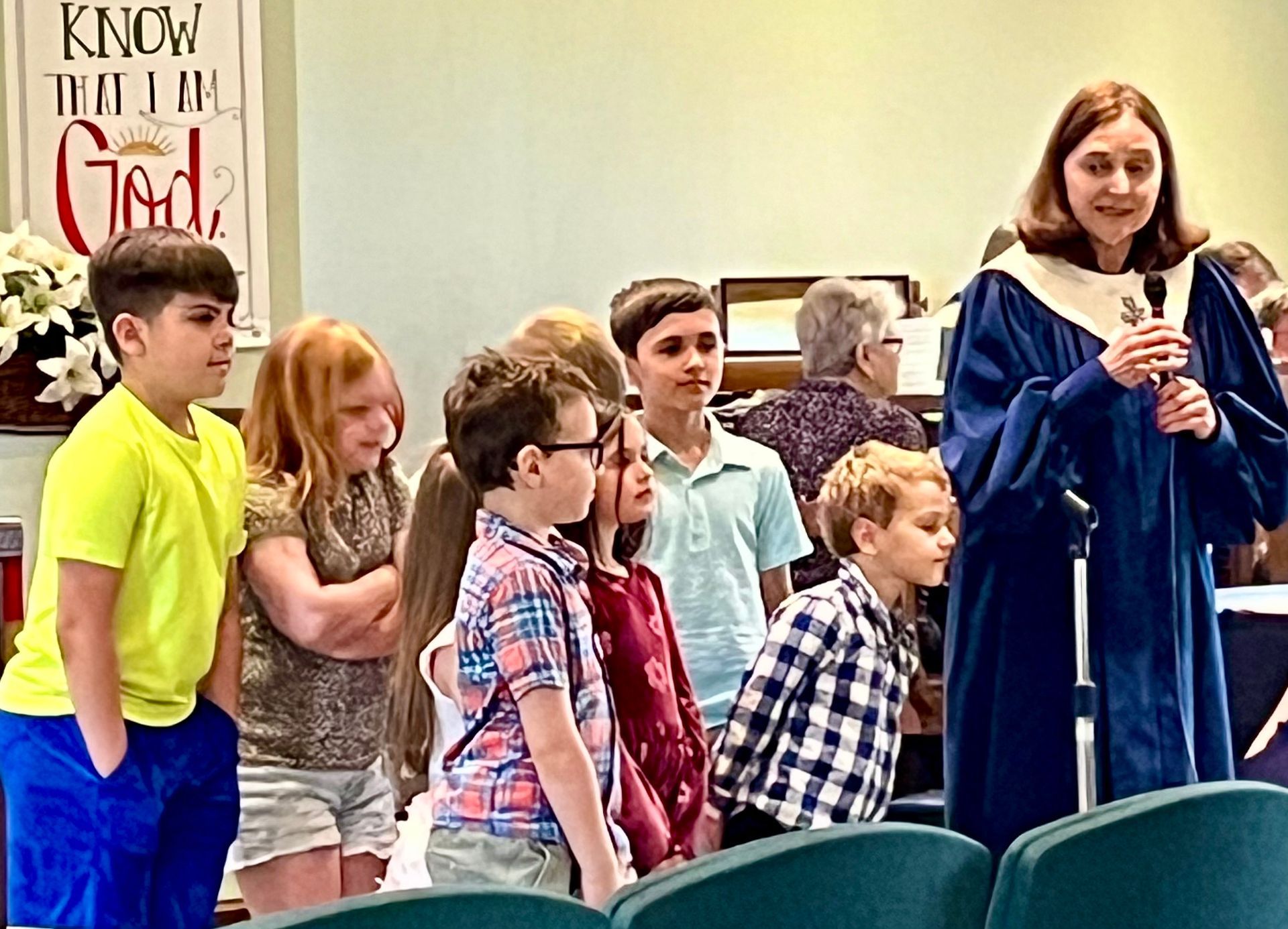 Woman in blue robe holding a microphone speaks to children standing in a church or assembly room.