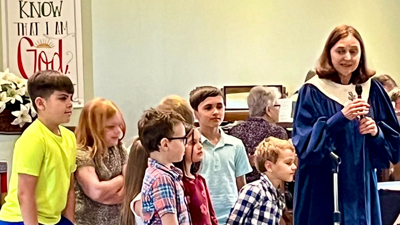 Woman in blue robe speaking to a group of children indoors near a “Know Him” poster.