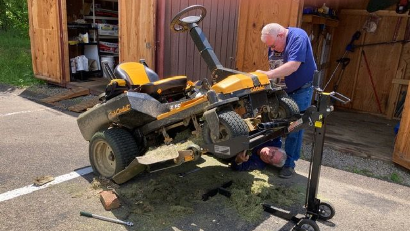 Two people repair a yellow riding lawn mower in a garage, one working underneath it.