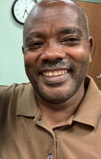 Smiling man in a brown polo shirt, seated indoors with a clock and wooden door behind him.