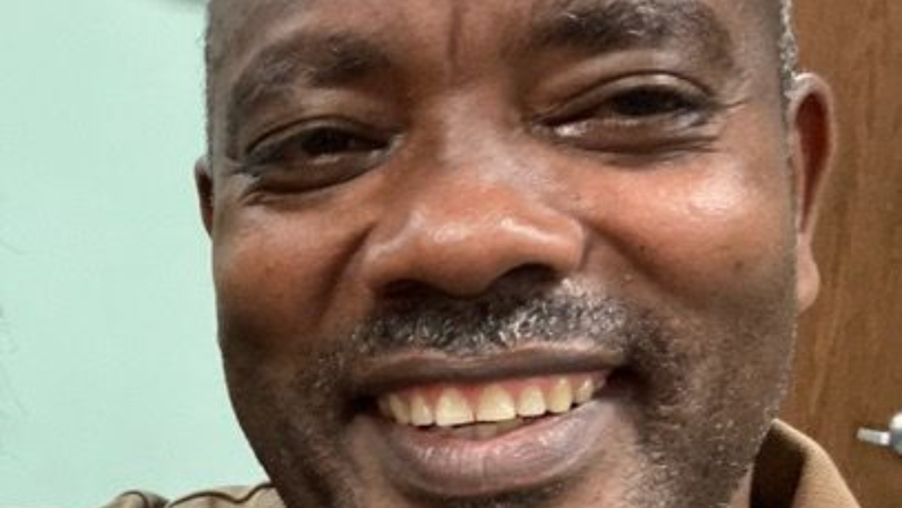 Close-up of a smiling man indoors against a light green wall and wooden door.