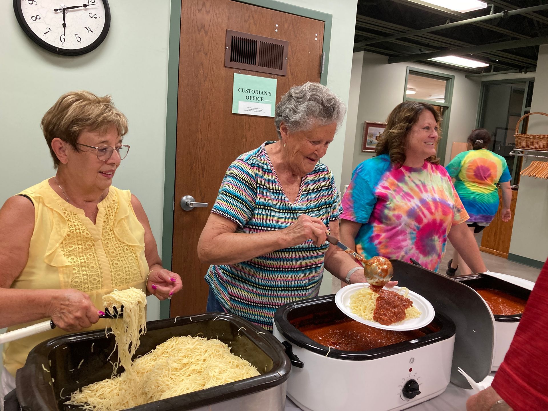 Three women serve food from slow cookers at a community buffet table.