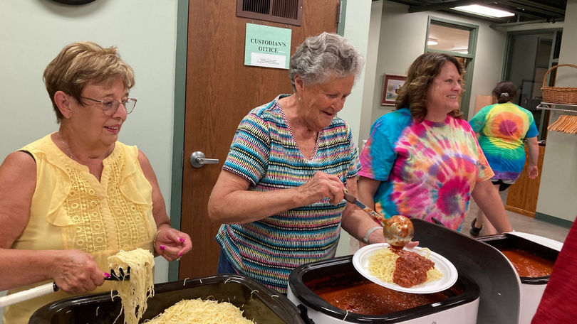 Three women serving food at a buffet table, one in a tie-dye shirt smiling at the counter