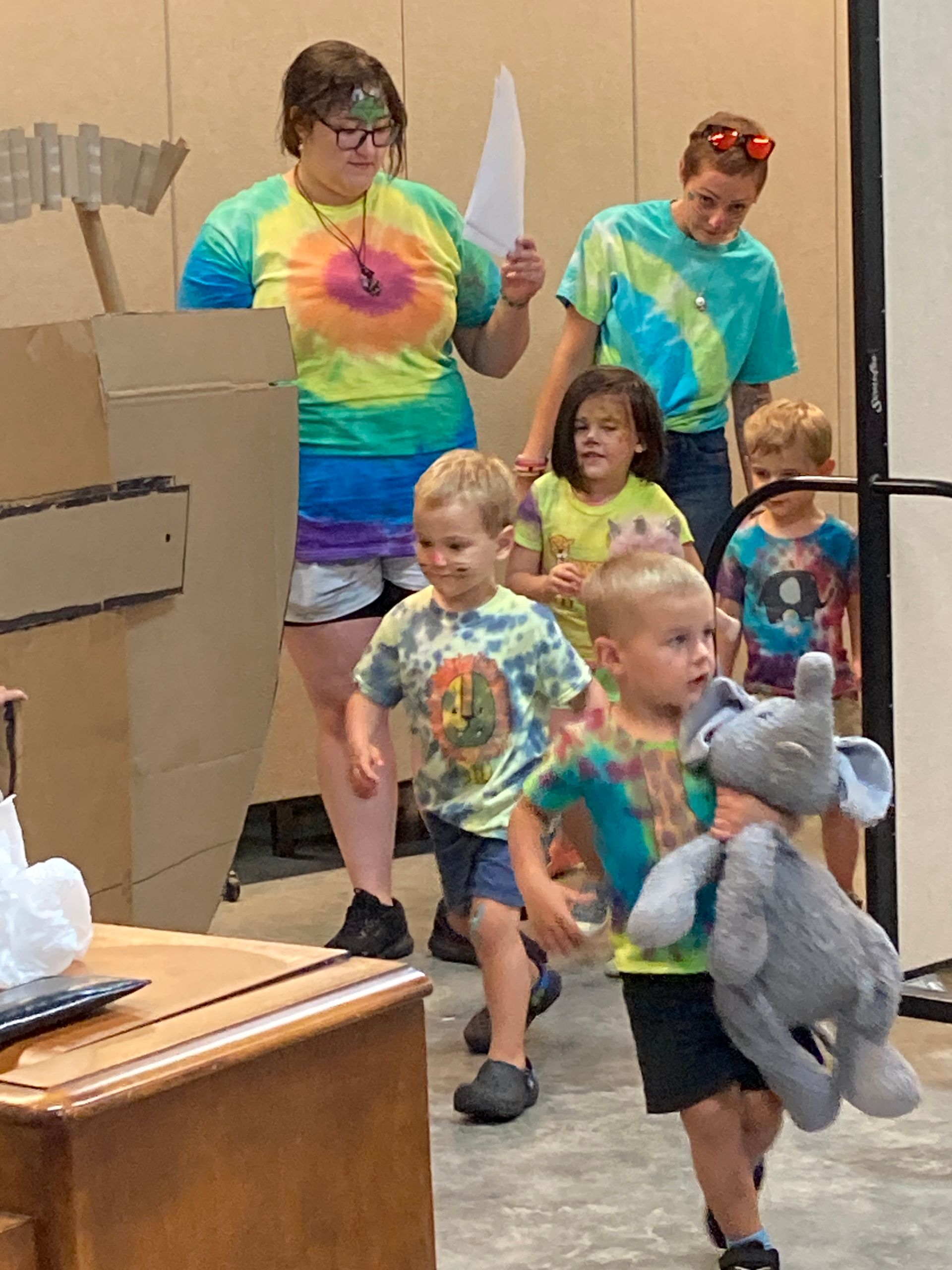 Children in tie-dye shirts march indoors, carrying cardboard swords and a stuffed elephant.