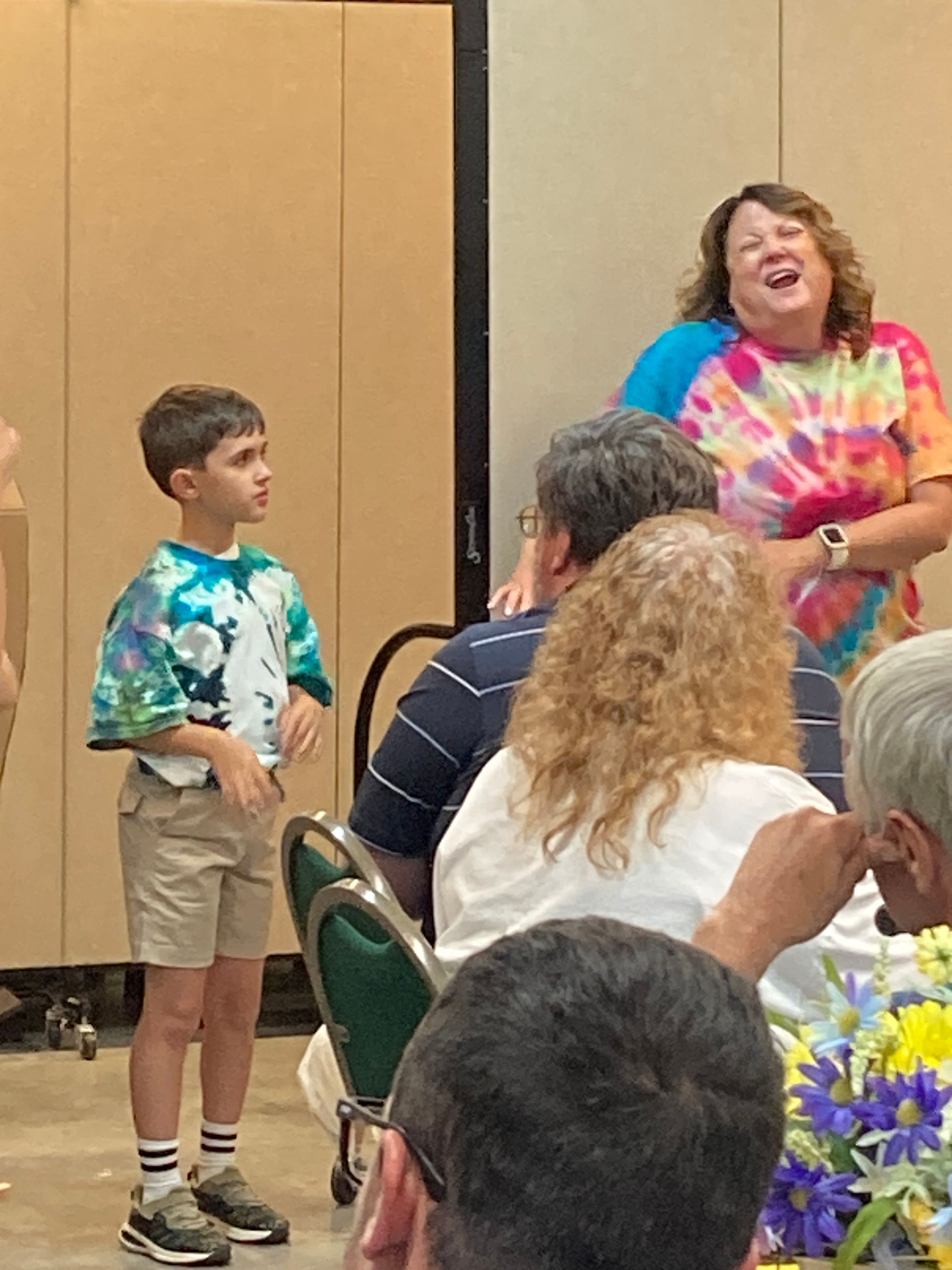 Child stands near a smiling speaker in a meeting room with seated attendees and flowers in the foreground