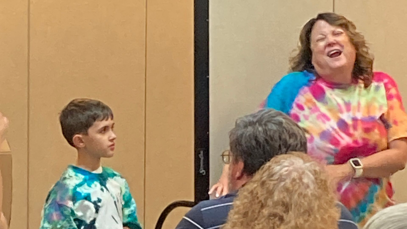 Woman in a tie-dye shirt speaking animatedly to a seated audience in a room with beige walls