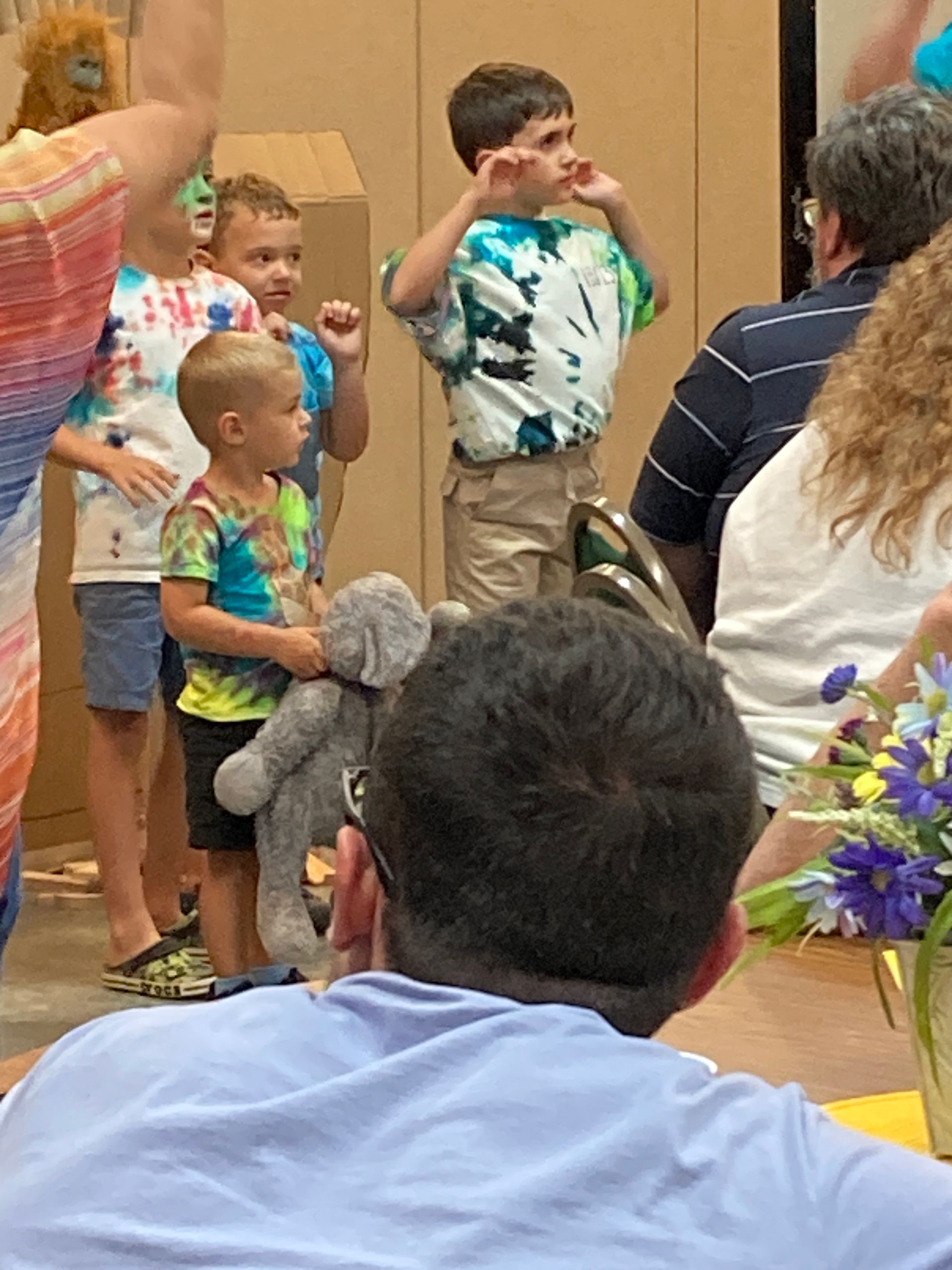 Children standing and sitting at a small indoor gathering, one boy in a tie-dye shirt looking to the side.