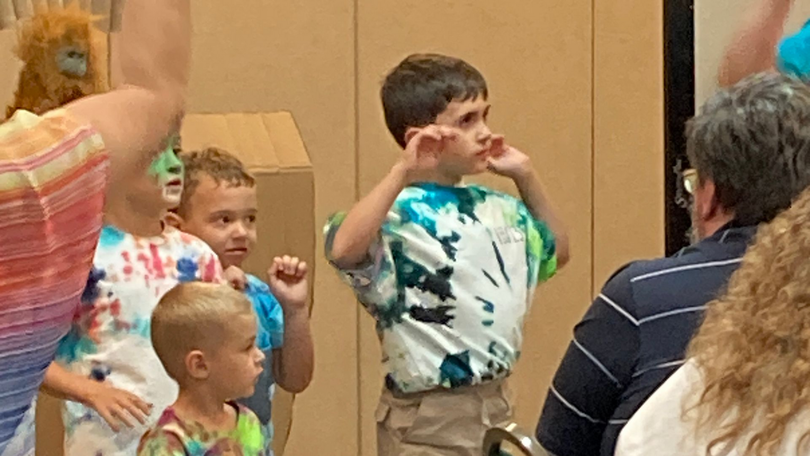 Children in tie-dye shirts making hand gestures in a room with an audience nearby