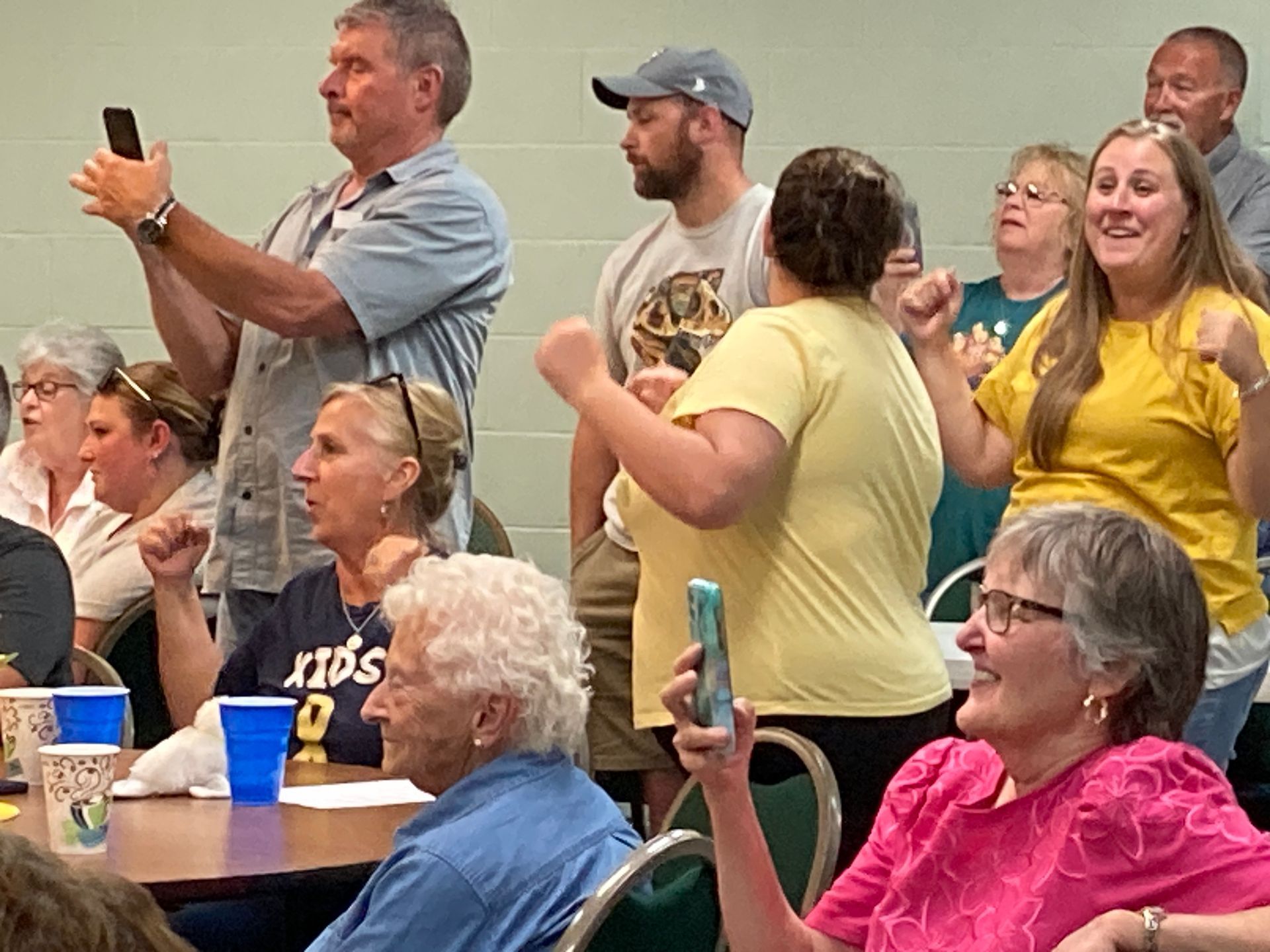Audience members standing and clapping in a meeting room, several holding up phones to record.