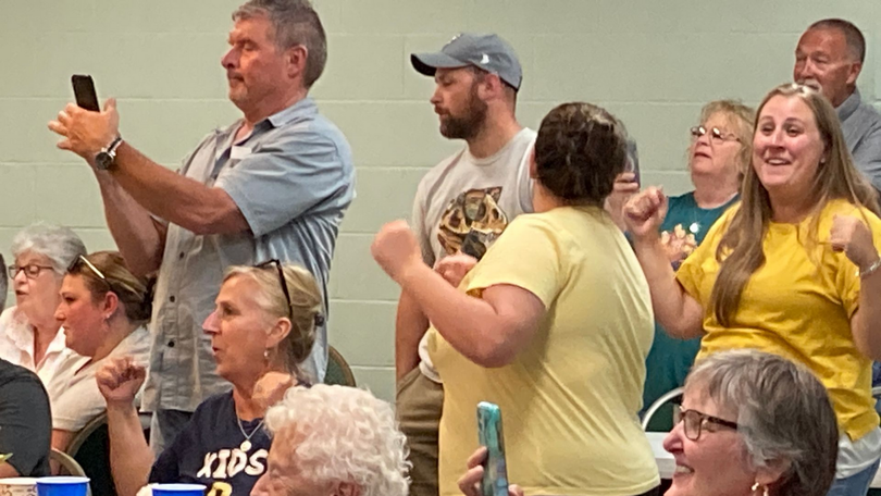 People standing and cheering in a crowded indoor event, with one man taking a phone photo.