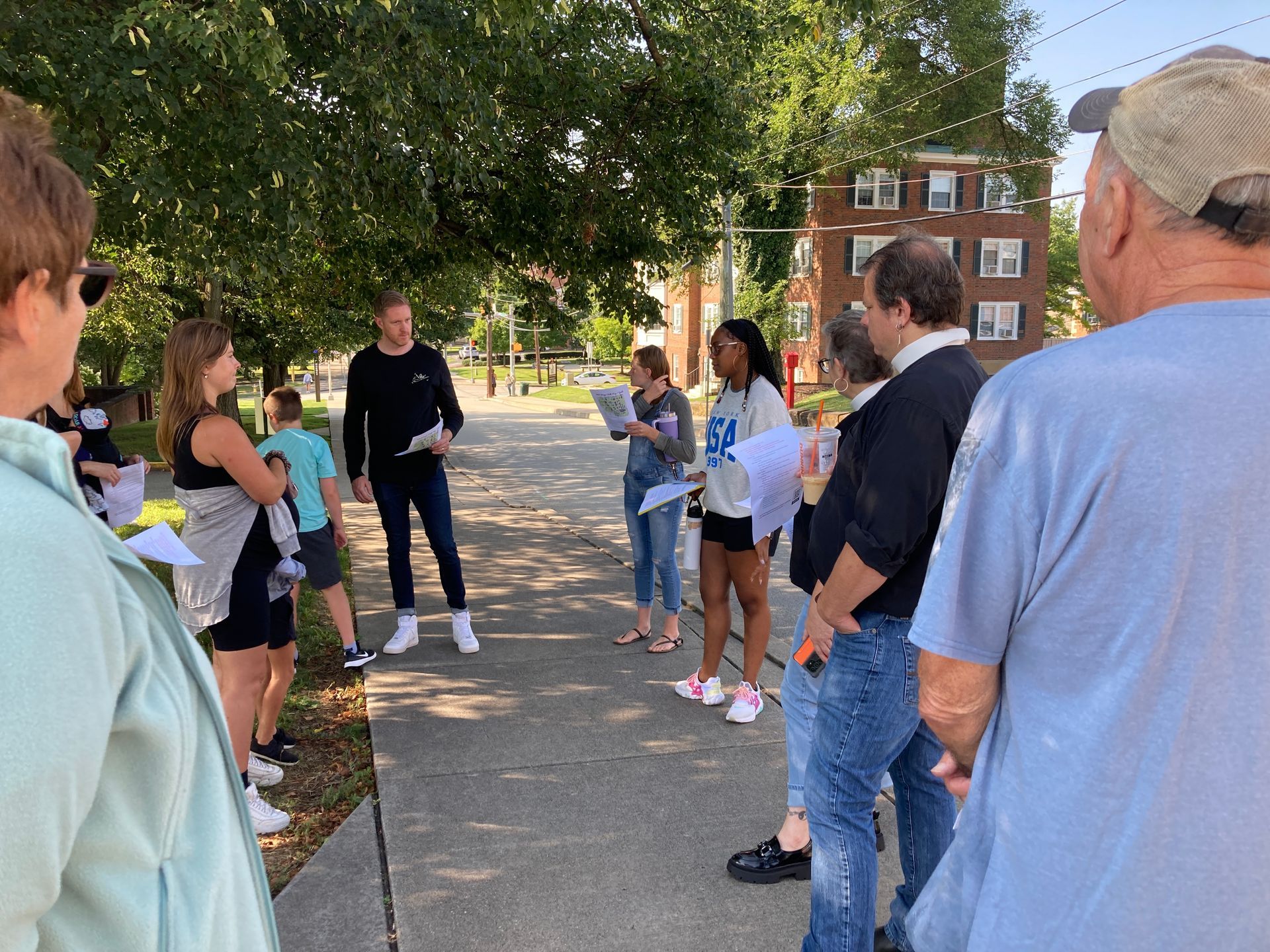 Group of people gathered outdoors on a sidewalk, listening to a speaker near a brick building and trees.
