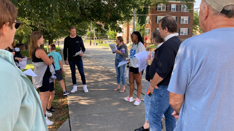 Group gathered on a shaded campus path, listening to a speaker near brick buildings and trees.