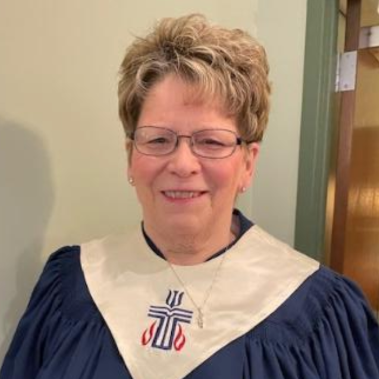 Smiling person in a blue choir robe with a white collar and cross emblem, standing indoors