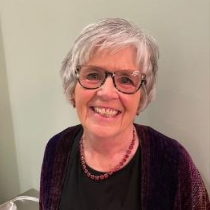 Smiling person with short gray hair, glasses, and a dark top against a plain wall