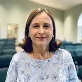 Woman smiling in a light blue embroidered blouse, seated indoors with a blurred auditorium background
