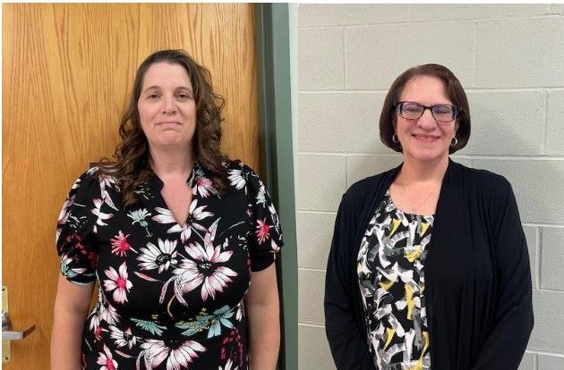 Two women standing indoors against a wall, one in a floral dress and one in glasses and a black cardigan.