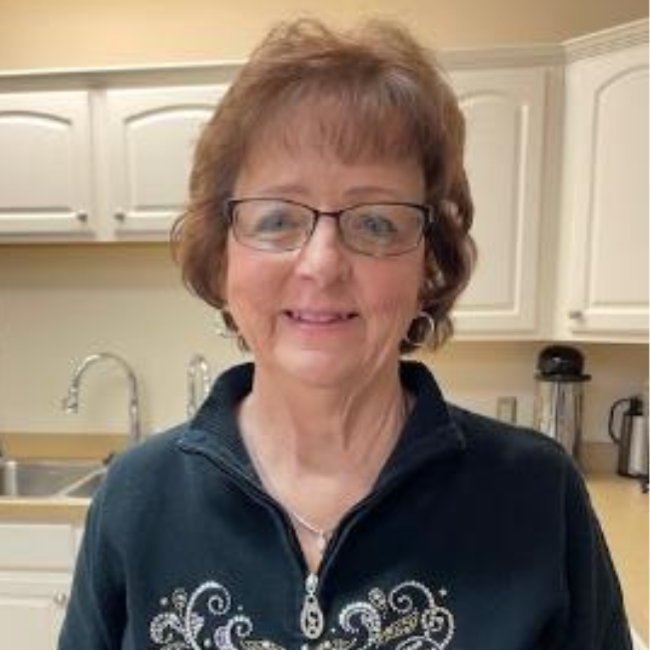 Woman smiling in a kitchen, wearing glasses and a dark zip-up top.