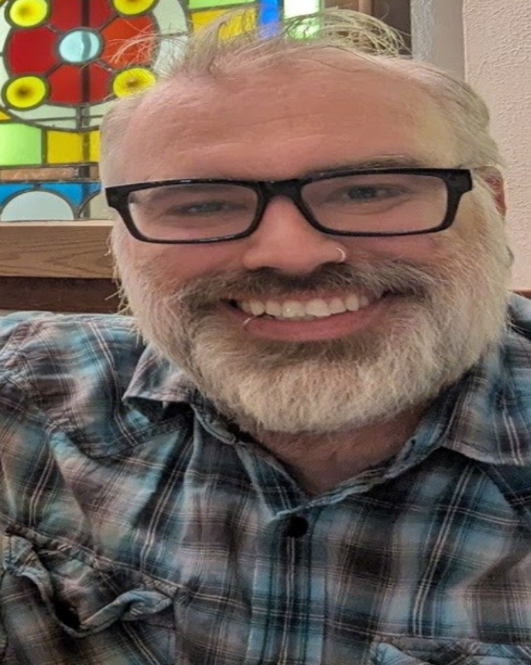 Smiling bearded man in black glasses and plaid shirt, seated indoors with a colorful stained-glass window behind him