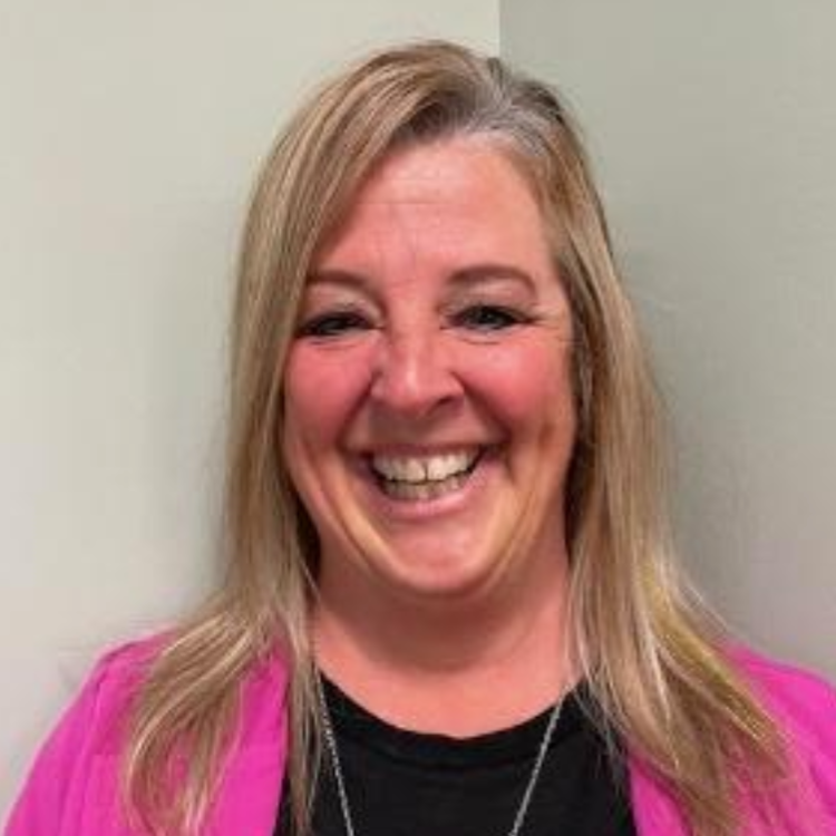 Smiling woman with blonde hair wearing a pink jacket and black top against a plain wall