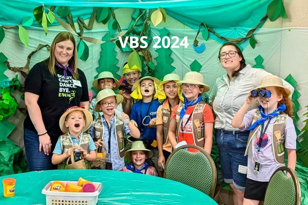 Group of children and adults posing under a green tent with “VBS 2024” banner and tropical decorations