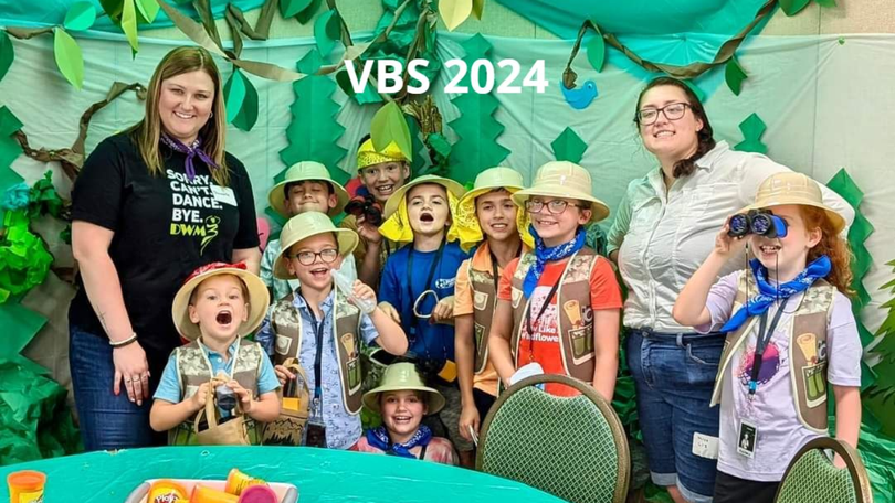 VBS 2024 group posing in leafy costumes and hats with two adults in front of a green backdrop