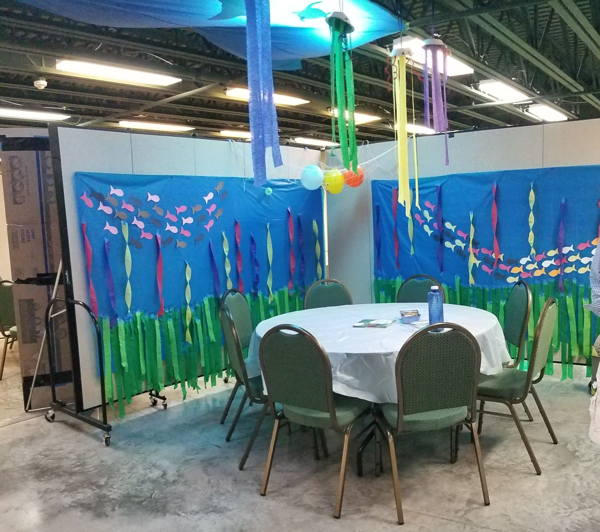 Decorated event table with green chairs in a blue, underwater-themed room with hanging streamers and balloons