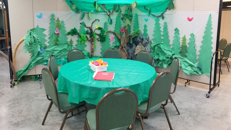 Round table with green tablecloth and chairs in a decorated outdoor party tent with forest backdrop