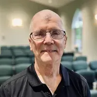 Smiling bald man wearing glasses and a black polo, seated indoors with blurred auditorium chairs behind him