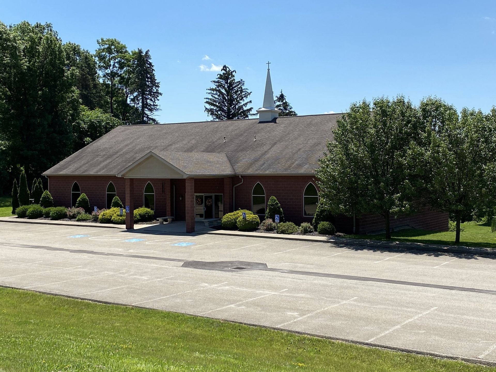 Small brick building with a shingled roof beside a parking lot, surrounded by trees and grass under a clear sky