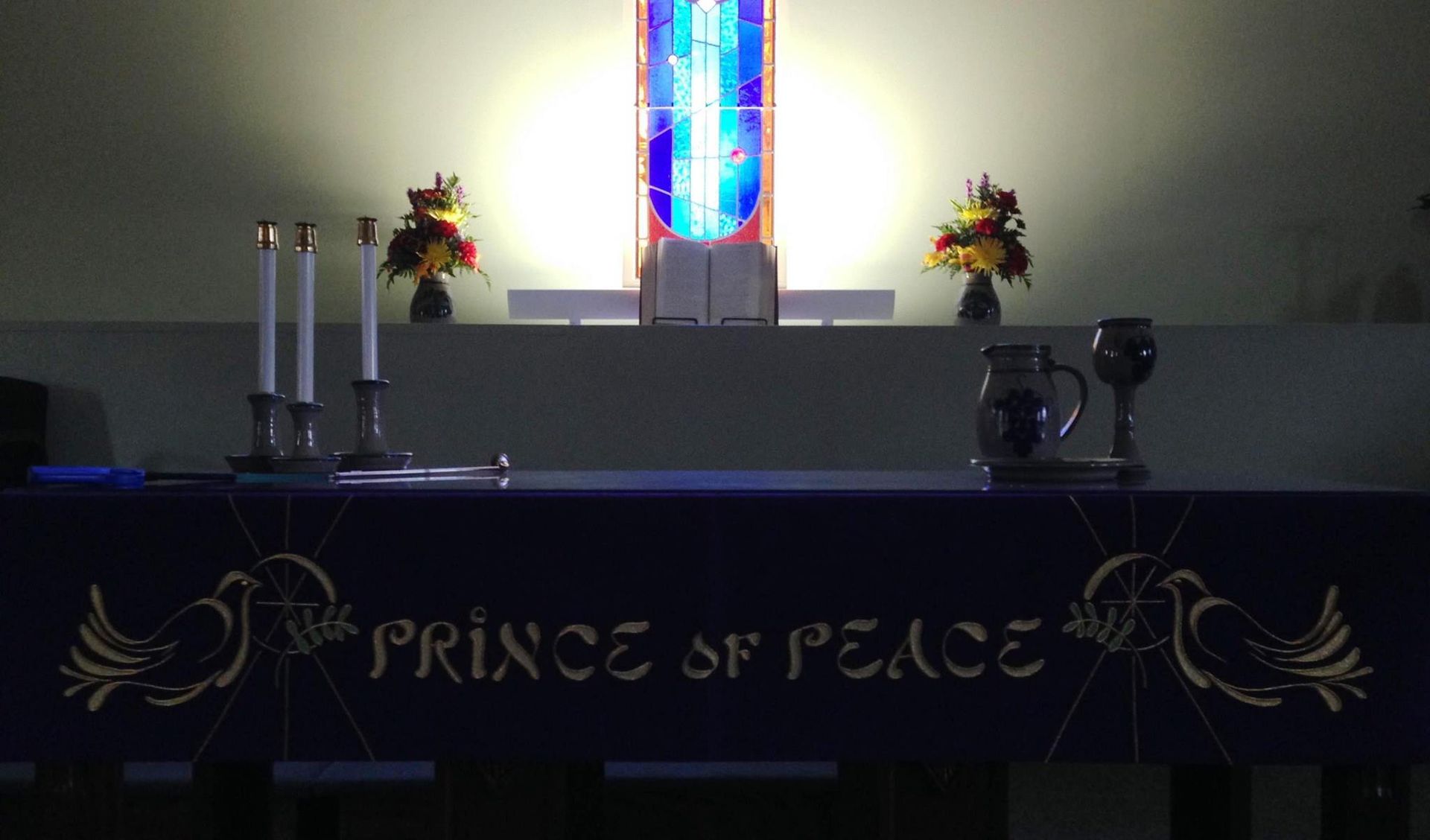 Church altar with colorful stained-glass cross, flowers, candles, and “Prince of Peace” banner
